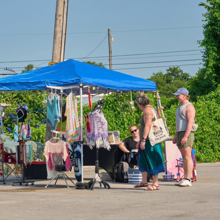 Shoppers browse handmade crochet apparel under a blue tent at the C-Street City Market.