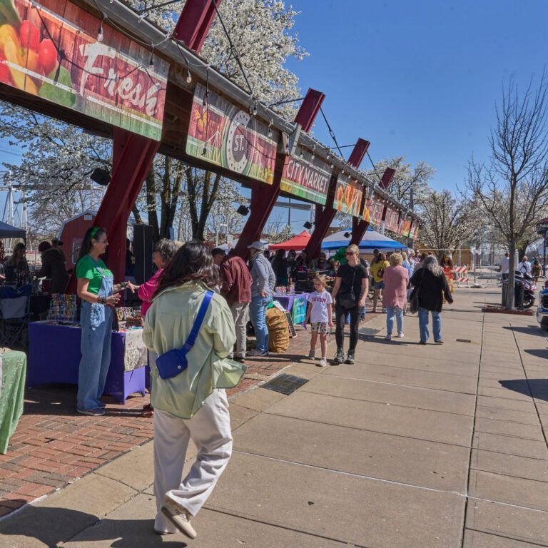 Shoppers walk beneath the Fresh City Market sign amid spring blossoms on Historic C-Street.