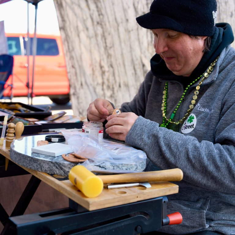 A leather artist at a C-Street market bench, wearing an "I love C-Street" sticker.