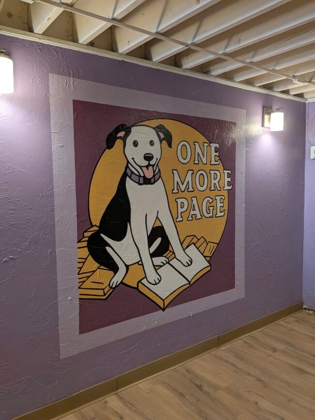 The One More Page dog mural, a black-and-white dog resting on an open book, painted on a purple wall.