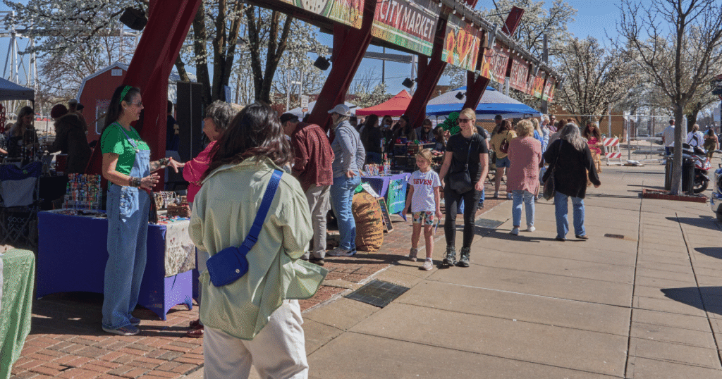 Shoppers and vendors under the red-beam pavilion at the C-Street City Market on a spring Saturday.