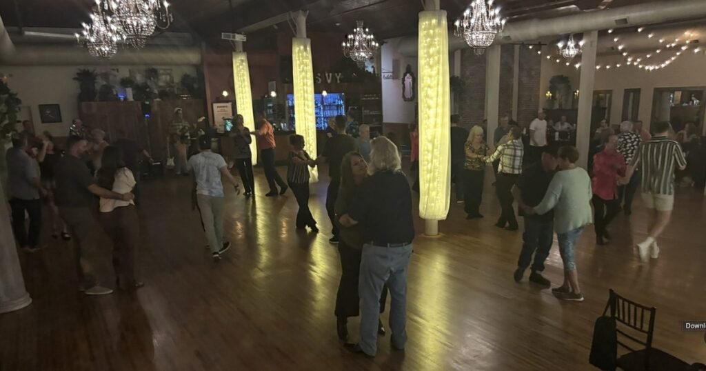 Couples dancing on a hardwood floor in a ballroom lit by crystal chandeliers, string lights, and glowing wrapped columns.