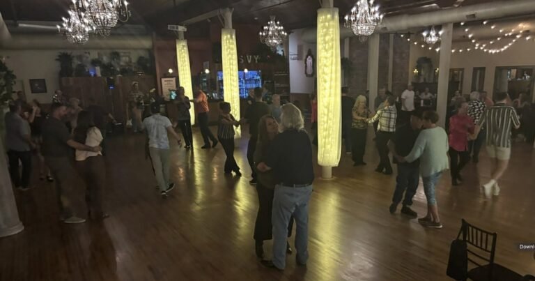 Couples dancing on a hardwood floor in a ballroom lit by crystal chandeliers, string lights, and glowing wrapped columns.
