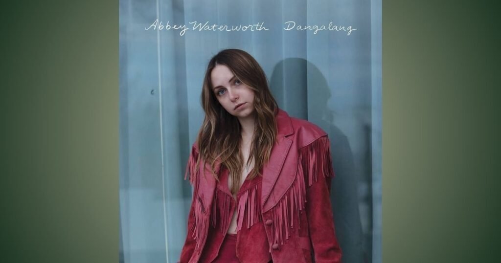 Abbey Waterworth in a red fringed suede jacket, leaning against a pale blue curtain with her head tilted.