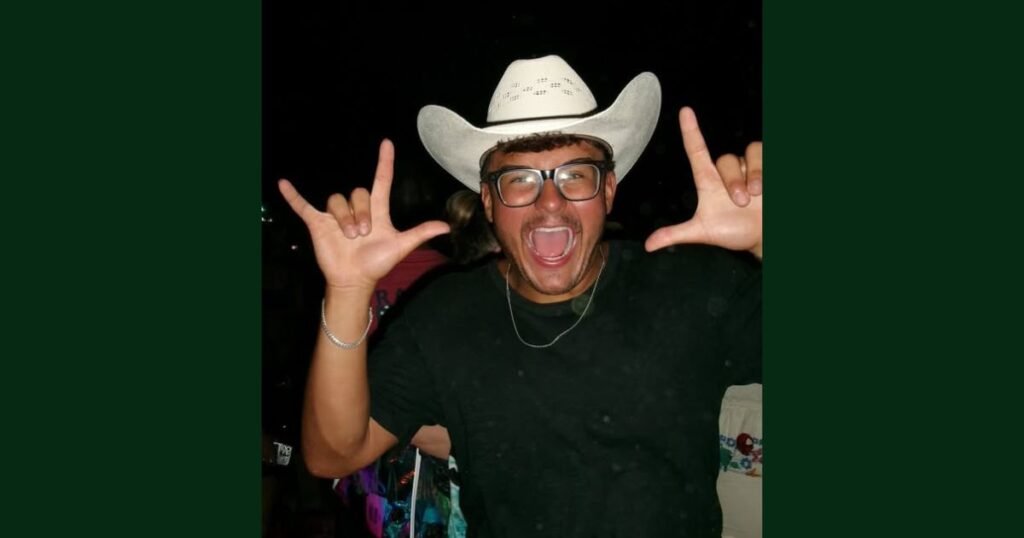 Riley Mays in a white cowboy hat and glasses, shouting with joy and throwing up rock-on hand signs with both hands.